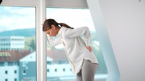 Junge-Frau-Verspürt-Rückenschmerzen-Vor-Dem-Fenster-In-Einem-Modernen-Apartmentbüro