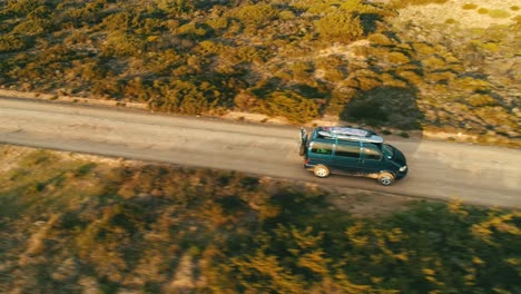 Old-nostalgic-van-bus-with-surfboards-on-roof-drives-along-dusty-road-in-Mediterranean-landscape