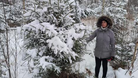 Woman-in-grey-furry-parka-carries-saw-and-shakes-snow-gently-off-of-conifer-tree