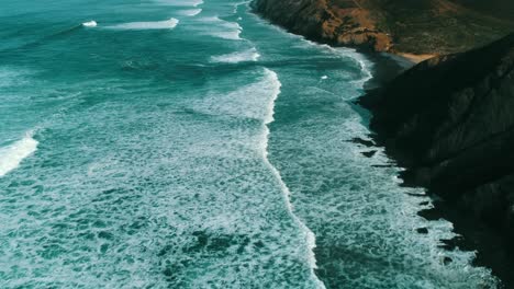 Restless-surf-of-blue-turquoise-ocean-meets-cliffs-and-sandy-bay-in-light-of-setting-sun,-drone-aerial-panorama