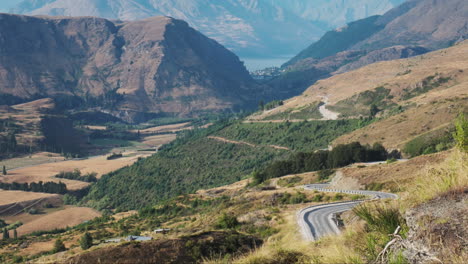 A-steep,-winding-mountain-road-carves-its-way-down-towards-the-vibrant-town-of-Queenstown,-New-Zealand