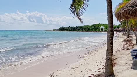 Langsamer-Schwenk-Nach-Links-An-Einem-Wunderschönen-Strand-In-Tulum-In-Der-Nähe-Von-Cancun,-Mexiko,-Mit-Kleinen-Wellen,-Palmen,-Blauem-Himmel-Und-Einigen-Wolken