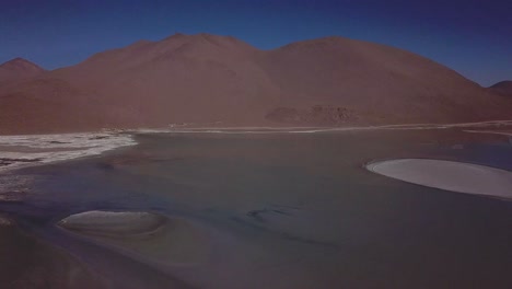 Die-Luftaufnahme-Zeigt-Die-Große-Höhenlage-Boliviens-Und-Zeigt-Eine-Trostlose-Landschaft,-In-Der-Der-Blick-Auf-Eine-Riesige-Wasseroberfläche-Gelenkt-Wird,-Die-An-Einen-Fernen-Berg-Angrenzt