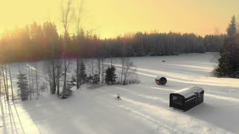 Aerial-view-flying-over-a-sauna-and-a-cabin-at-a-forest-lake,-during-sunset