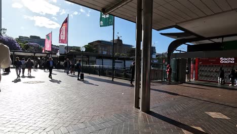 People-walking-through-Circular-Quay,-Sydney-on-sunny-day-with-flags-blowing-in-the-breeze