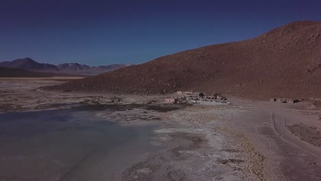 Über-Dem-Hochgelegenen-Plateau-Boliviens-Schwebend,-Enthüllt-Die-Luftaufnahme-Eine-Trostlose-Landschaft,-In-Der-Die-Weite-Des-Geländes-Durch-Eine-Kleine-Dorfsiedlung-Unterbrochen-Wird
