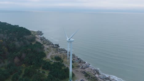 Establishing-aerial-view-of-abandoned-seaside-fortification-buildings-at-Karosta-Northern-Forts-on-the-beach-of-Baltic-sea-in-Liepaja,-overcast-day,-wind-turbine,-drone-orbit-shot