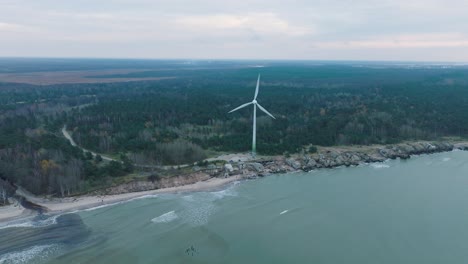 Establishing-aerial-view-of-abandoned-seaside-fortification-buildings-at-Karosta-Northern-Forts-on-the-beach-of-Baltic-sea-in-Liepaja,-overcast-day,-wind-turbine,-drone-orbit-shot