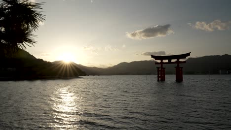 Silhouette-Des-Großen-Torii-Tors-Von-Itsukushima,-Das-In-Der-Bucht-Von-Hiroshima-Schwimmt,-Mit-Sonnenuntergangsfackeln-Im-Hintergrund-über-Der-Bergkette