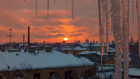 Orange,-glowing-sunset-over-a-snowy-village-in-winter-with-icicles-in-the-forground