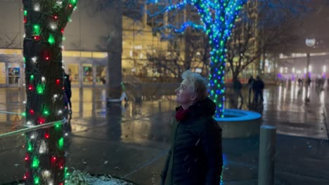 Ältere-Frau-Spaziert-Durch-Eine-Stadt,-Die-Mit-Weihnachtsbeleuchtung-Und-Nacht-Sowie-Schnee-Geschmückt-Ist