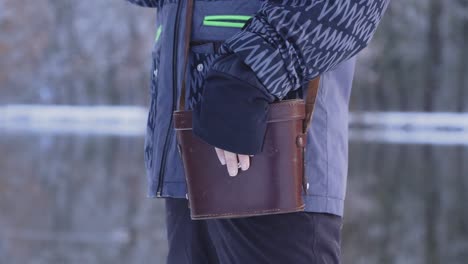 A-person-in-a-blue-jacket-standing-on-a-snowy-riverbank-has-his-hand-on-a-leather-case-for-binoculars