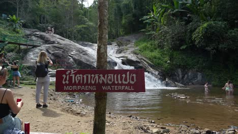 View-of-a-tourist-standing-by-the-sign-with-the-name-of-the-Mo-Pang-waterfall,-with-a-green-forest-in-the-background