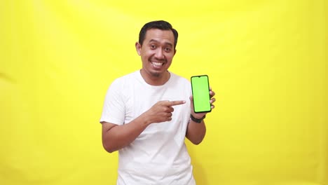 Asian-adult-man-showing-green-screen-mobile-phone-by-pointing-finger-while-looking-camera-isolated-on-yellow-background---concept-of-advertisement