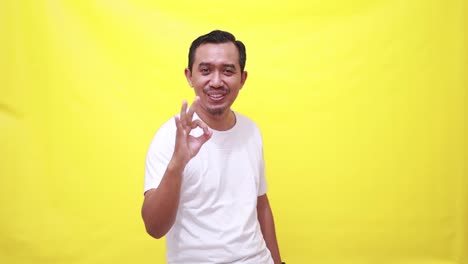 Handsome-asian-man-showing-okay-gesture-with-one-hand-while-looking-at-camera-and-smiling-while-standing-against-yellow-background