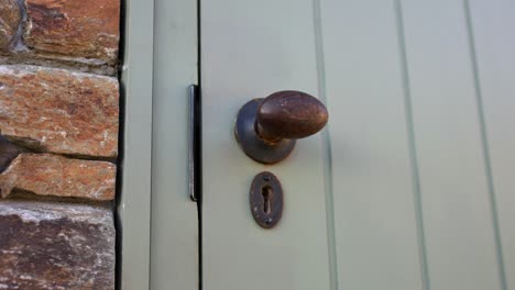 Close-up-of-old-fashioned-brass-door-handle-into-a-rustic-farmhouse-building-with-light-painted-wooden-door-and-stone-work