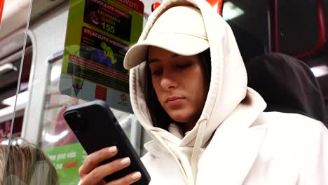 Woman-in-white-hoodie-and-cap-meaninglessly-scroll-phone-while-in-metro-wagon