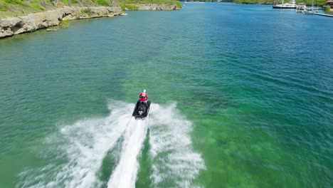 Rear-view-tracking-follows-man-standing-up-on-jetski-racing-over-green-blue-Caribbean-water,-aerial