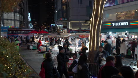 La-Gente-Que-Viaja-Come-Comida-Callejera-En-El-Mercado-Nocturno-De-Myeongdong-En-Seúl,-La-Calle-Comercial-Del-Centro.