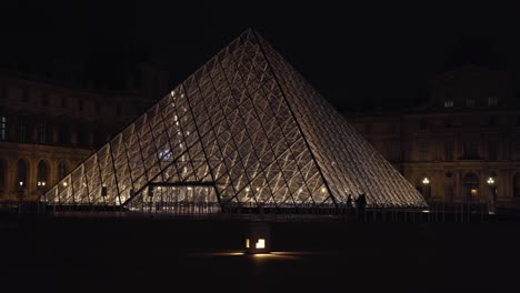 Pirámide-Del-Louvre-Por-La-Noche-Con-Siluetas-De-Personas-Delante-De-Ella.