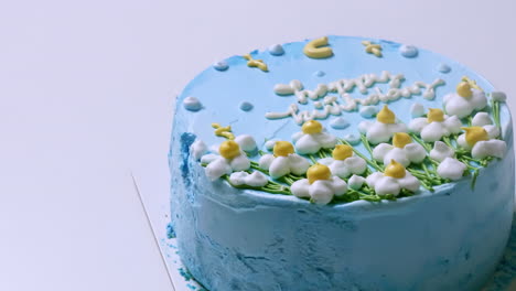 Close-up-shot-of-the-top-of-a-birthday-cake-with-blue-frosting-and-decorated-with-white-and-yellow-frosted-flowers,-with-happy-birthday-greetings-on-top-of-it