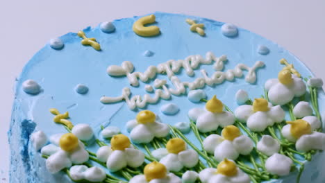 Zooming-out-of-a-close-up-shot-of-a-blue-frosted-birthday-cake,-decorated-with-small-white-flowers-with-yellow-tips-and-green-accents