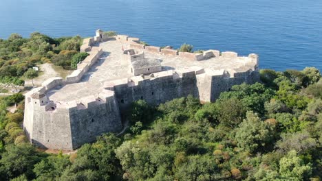 Drone-view-in-Albania-flying-around-a-medieval-fort-on-a-green-island-over-blue-clear-ocean-on-a-sunny-day