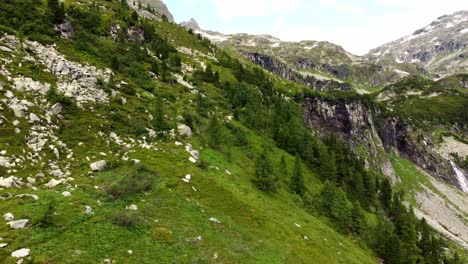 Steep-slope-covered-in-grass-with-a-lot-of-stones-next-to-a-huge-cliff-and-a-large-waterfall-in-the-Alps-in-Kaernten,-Austria