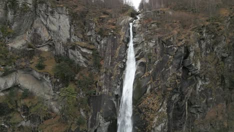 Retreating-drone-shot-of-a-light-snowfall-in-front-of-a-cascading-Cascata-di-Foroglio-Waterfall,-situated-in-the-village-of-Cavergno,-in-the-district-of-Vallemaggia,-in-Switzerland