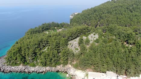 Drone-view-in-Albania-flying-over-a-green-forest-next-to-a-blue-ocean-water-on-a-sunny-day