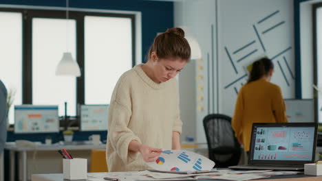 Busy-woman-in-startup-office-holding-clipboard-looking-at-papers-with-sales-charts