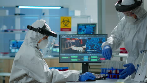 Biologist-doctor-in-ppe-suit-checking-liquid-sample-typing-on-pc
