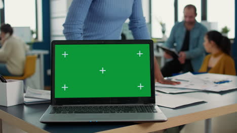 Laptop-computer-with-green-screen-on-desk-in-front-of-african-american-startup-employee-looking-at-paper