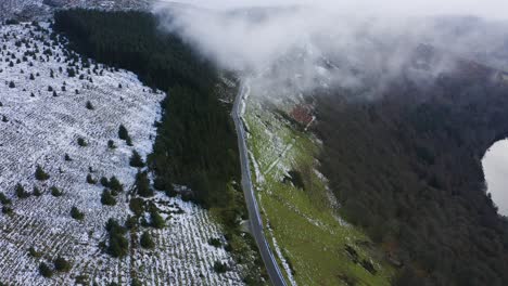 Winter-Tranquility:-Aerial-View-of-an-Empty-Wicklow-Mountain-Road-Split-by-Snow,-Enveloped-in-Low-Clouds,-Revealing-the-Serene-Beauty-of-Forests-and-Guinness-Lake