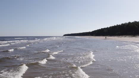 Drone-dolly-above-waves-crashing-on-shallow-sandy-beach-as-lone-man-walks-on-coast
