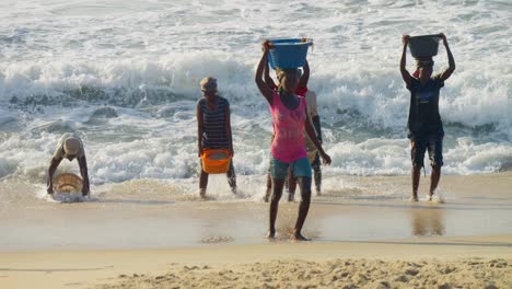 African-women-carrying-baskets-of-sand-on-heads,-walk-out-of-sea-waves