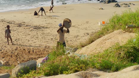 African-woman-emptying-bucket-of-sand-on-beach,-kids-playing-around