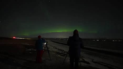 Statische-Aufnahme,-Die-Die-Silhouette-Zweier-Fotografen-Zeigt,-Die-über-Ein-Bild-Von-Grünen-Nordlichtern-Am-Dunklen-Himmel-In-Island-Sprechen