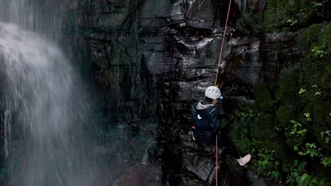 Aerial-view-of-female-climber-going-down-sheer-mountain-face-wall-beside-waterfall