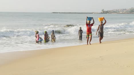 Afrikanische-Frauen-Tragen-Körbe-Auf-Dem-Kopf-Am-Strand,-Andere-Warten-In-Wellen