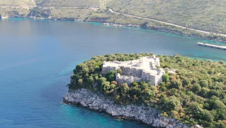 Drone-view-in-Albania-flying-away-from-a-green-island-with-a-medieval-fort-on-it-on-blue-clear-ocean-water-on-a-sunny-day