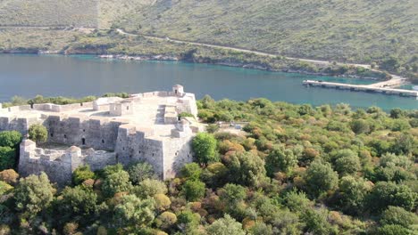 Drone-view-in-Albania-flying-into-a-green-island-with-a-medieval-fort-on-it-on-blue-clear-ocean-water-on-a-sunny-day