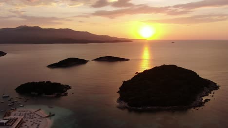 Drone-view-in-Albania-flying-over-blue-crystal-clear-water-on-sunset,-with-the-sun-setting-in-the-horizon-with-small-islands-in-Ksamil