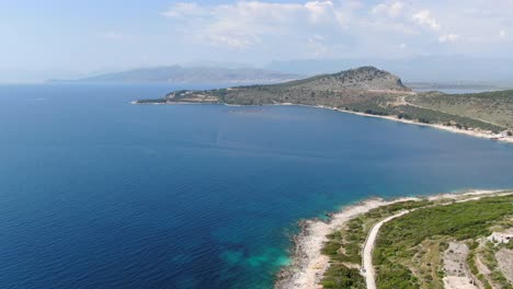 Drone-view-in-Albania-flying-over-blue-water-sea-next-to-green-mountains-landscape,-sunny-day-in-Ksamil