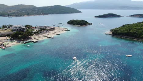 Drone-view-in-Albania-flying-over-white-sand-beaches,-blue-crystal-clear-water,-green-small-islands,-boats-and-hotels,-sunny-day-in-Ksamil