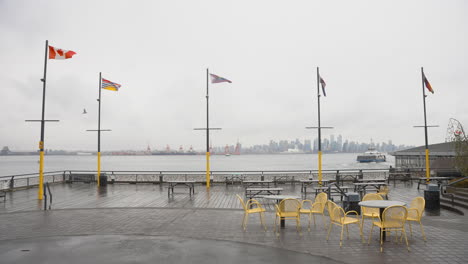 Un-Grupo-De-Sillas-Amarillas-Se-Encuentra-Vacía-En-Un-Muelle-Contra-El-Horizonte-De-La-Ciudad-De-Vancouver,-Con-El-Seabus-Y-Varias-Banderas-Visibles.