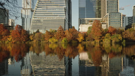 Beautiful-trees-and-downtown-city-buildings-with-reflection-in-water-from-lake-during-autumn-fall-sunset-with-pretty-colors-in-tree-leaves