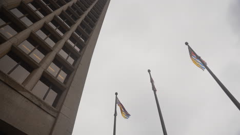 An-orbiting-shot-of-Canadian-and-British-Columbia-flags-near-a-tall-skyscraper-with-a-cloudy-backdrop