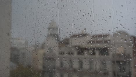 Eine-Nahaufnahme-In-Zeitlupe-Von-Schweren-Regentropfen,-Die-Auf-Einer-Fensterscheibe-Landen,-Mit-Einer-Städtischen-Stadtlandschaft-Im-Hintergrund