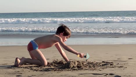 Young-boy-playing-on-the-beach-with-toy-shovel-and-making-sand-castle-while-people-walk-and-enjoy-the-beach-view